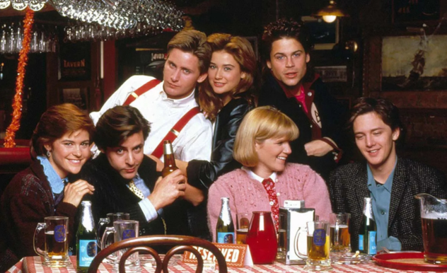 The cast is seated together at a table by the bar - the table littered with various beverages as the all pose looking at a different member of the cast.