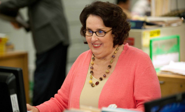 Phyllis, in a pink cardigan, sits at her desk and smiles at her computer screen.