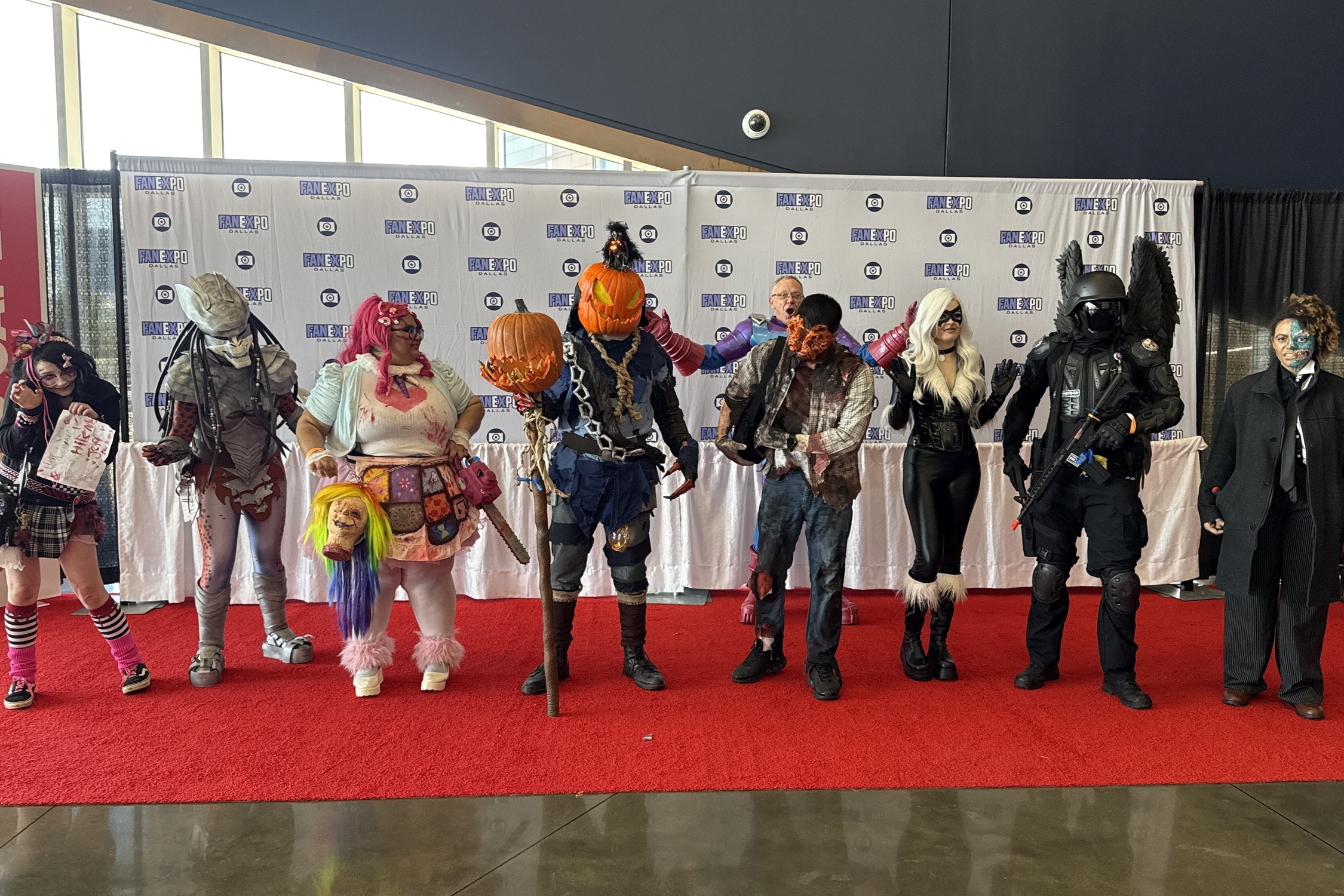 A lineup of cosplayers pose on the red carpet in front of a FAN EXPO Dallas step-and-repeat backdrop, showcasing elaborate costumes including horror, sci-fi, and fantasy designs such as a Predator, a pumpkin-headed scarecrow, a zombie, and a winged armored soldier.