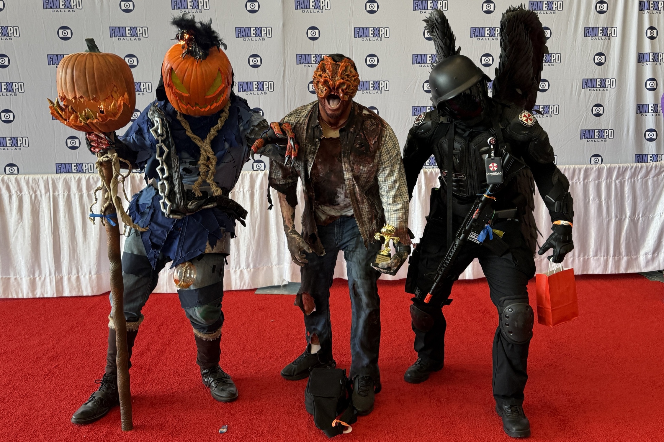 Three cosplayers stand together on the red carpet — one dressed as a pumpkin-headed scarecrow holding a staff, another as a grotesque zombie with a trophy in hand, and the third in full tactical armor with black wings and a replica rifle.
