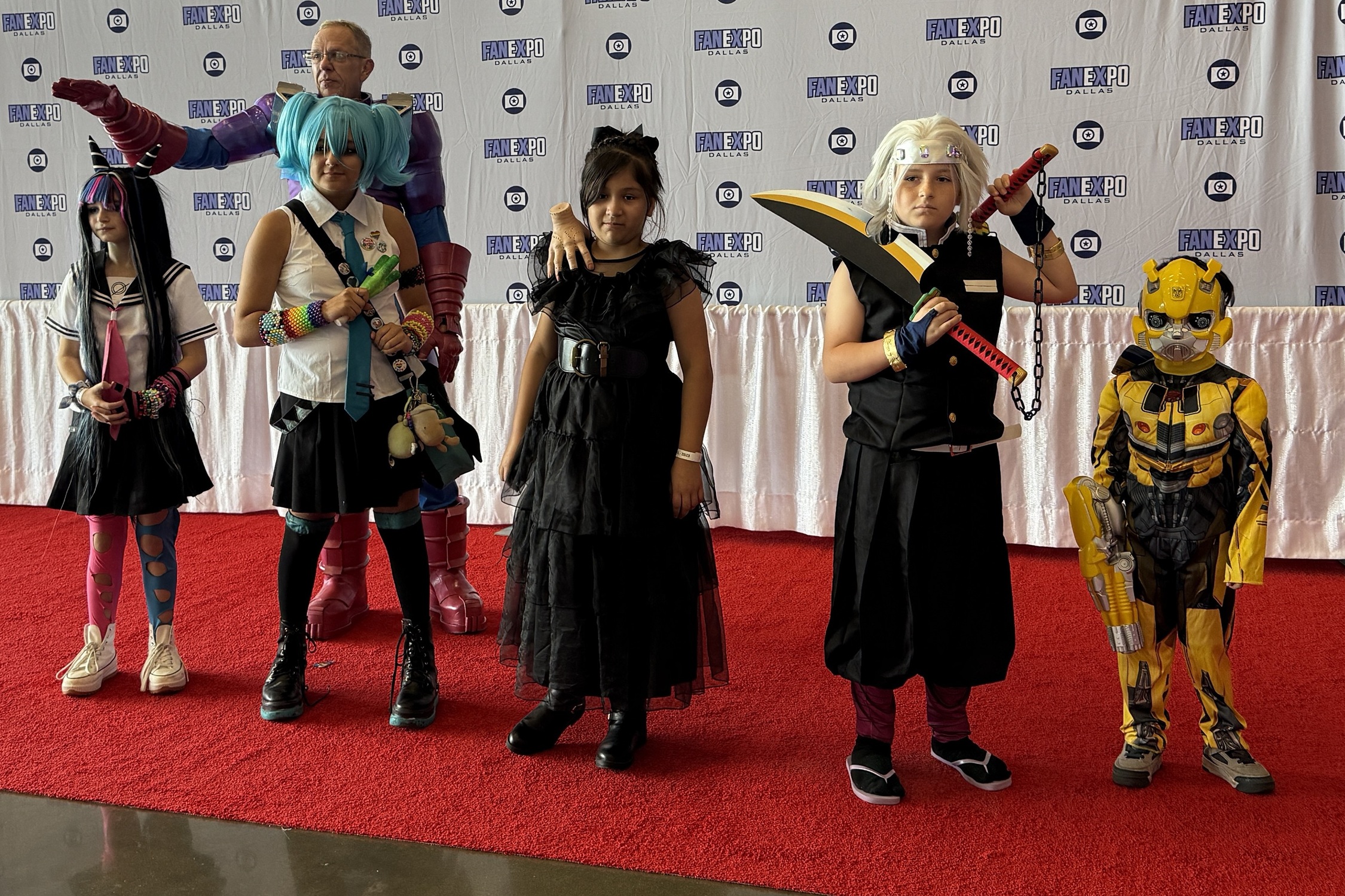Young cosplayers stand proudly on the red carpet in front of a FAN EXPO Dallas backdrop, dressed as a mix of anime, superhero, and fantasy characters, including Bumblebee, Mitsuri Kanroji, and others, with an adult cosplayer in a towering Galactus costume standing behind them.