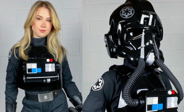 Cosplayer poses against a grey brick background and is wearing a starfighter pilot uniform with a black helmet.