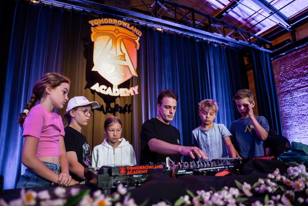 Group of children learning DJ skills from an instructor at Tomorrowland Academy, standing around a DJ console with the Tomorrowland Academy banner in the background.
