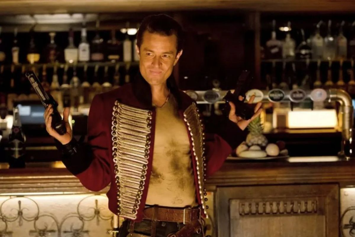 In a old military uniform, holding two guns, James Marsters is standing in front of a bar
