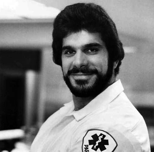 Black and white image of Lou Ferrigno smiling at the camera, wearing a white paramedic uniform with a patch on his sleeves.