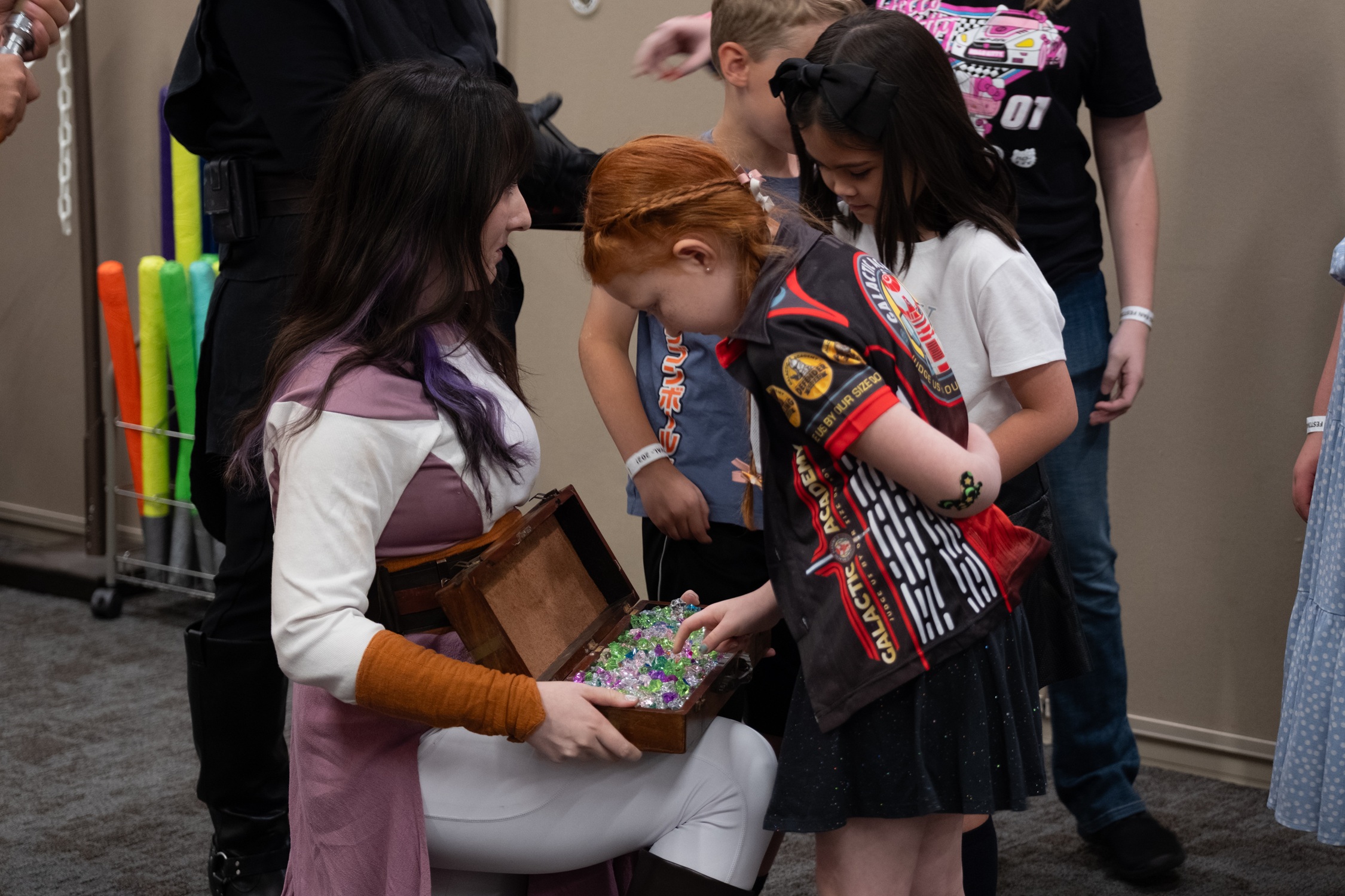 A cosplayer dressed as a Star Wars character kneels and opens a small treasure chest filled with colorful crystals for a group of excited kids.