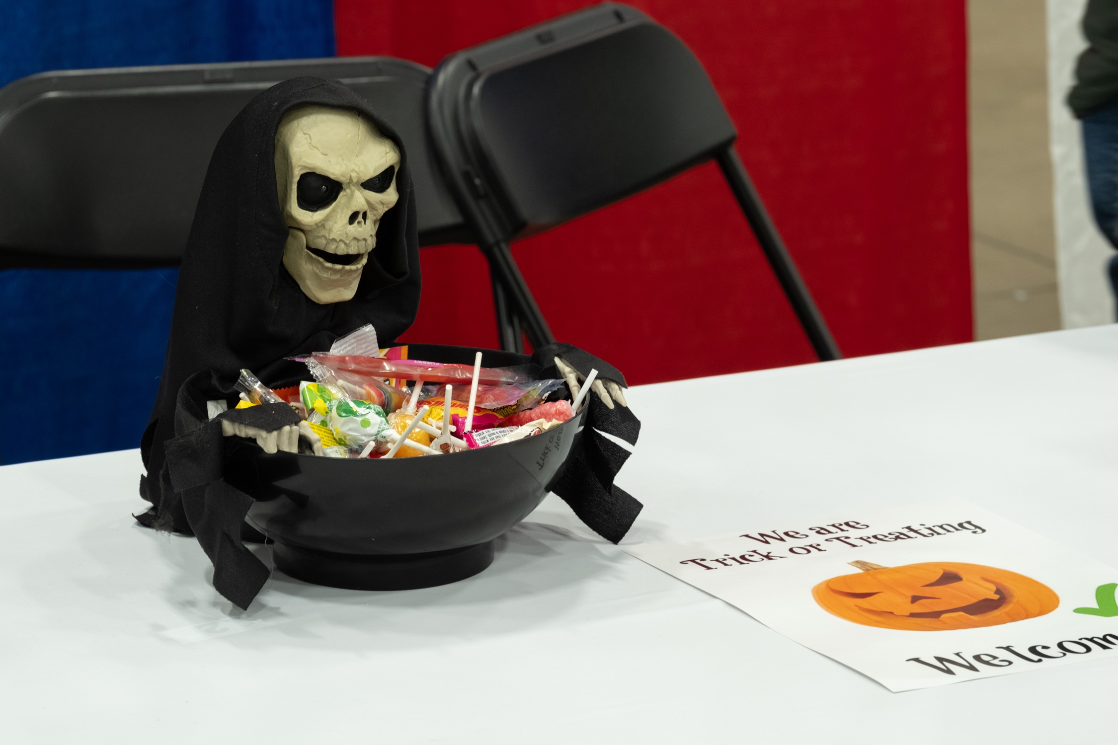 A Halloween candy bowl decorated with a skeleton in a black cloak sits on a table beside a sign that reads “We are Trick or Treating – Welcome!” with a smiling jack-o’-lantern graphic.