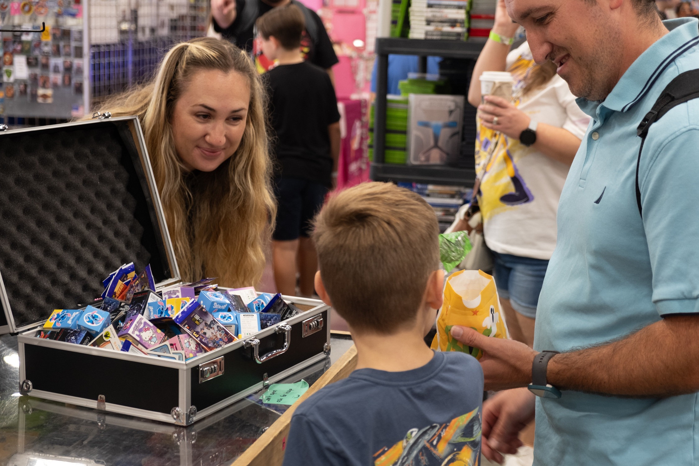 A vendor smiles as a young boy with his dad approaches a table filled with colorful treat boxes, collecting candy during trick-or-treating on the show floor.