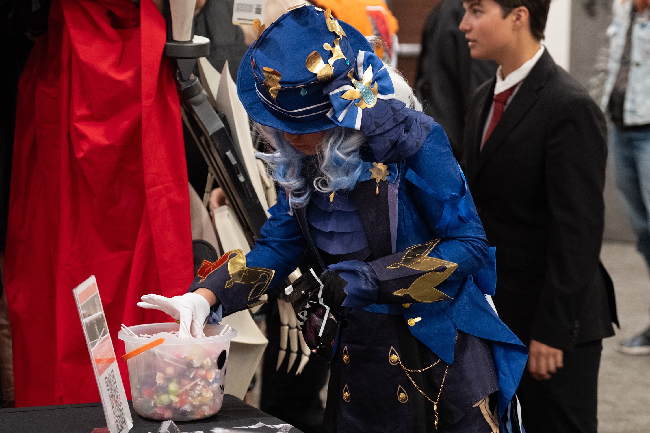 A cosplayer dressed in an elaborate blue and gold costume with a wide-brimmed hat selects candy from a treat bowl at a booth.