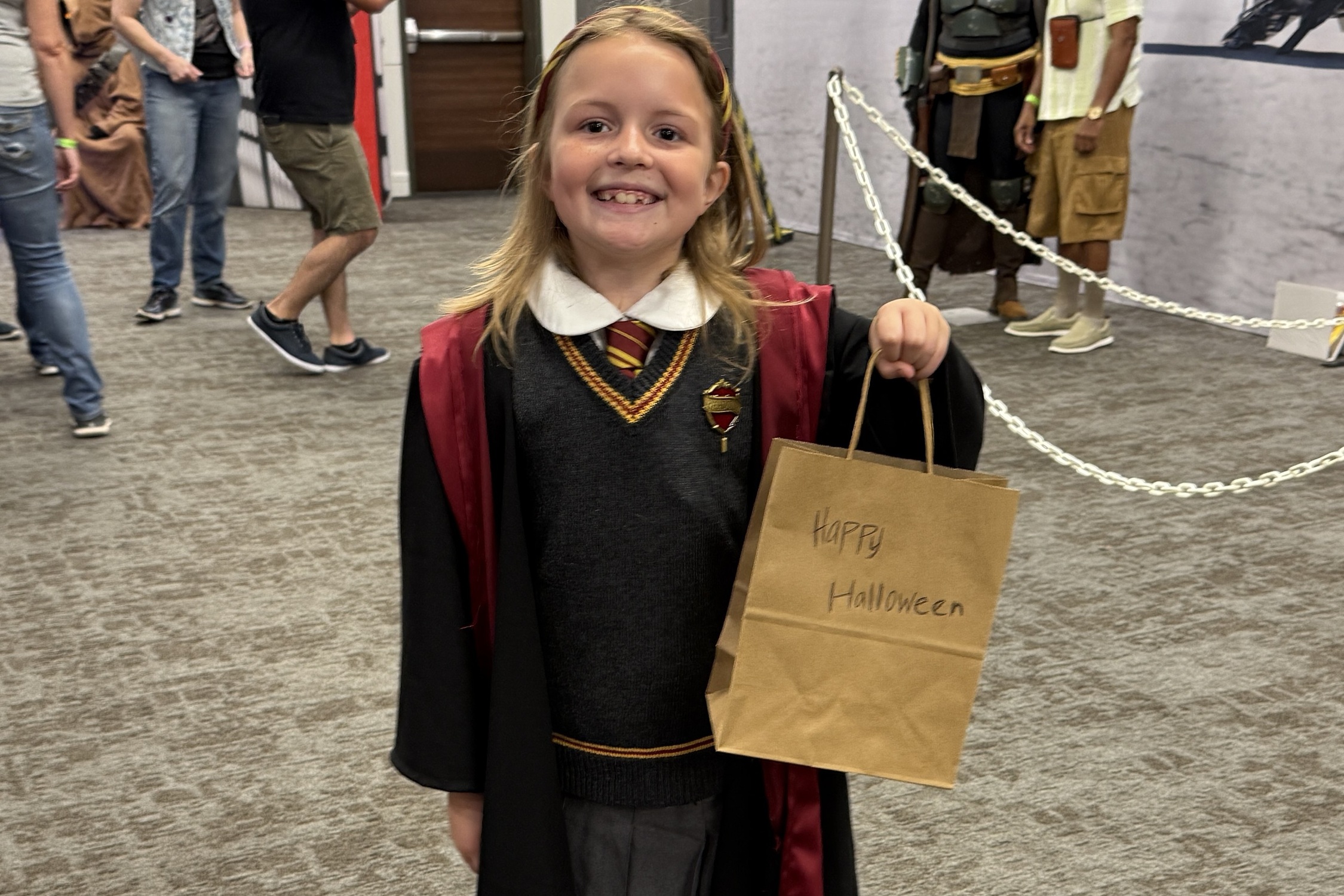 A smiling child dressed as a Hogwarts student from Harry Potter holds up a brown paper bag labeled “Happy Halloween.”