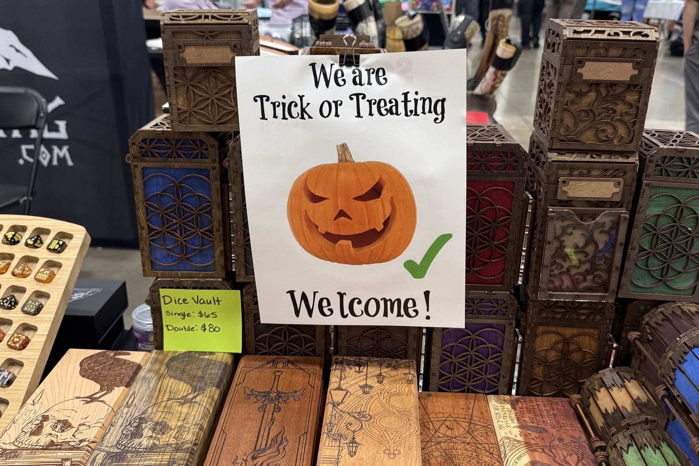 A sign with a carved pumpkin graphic and green checkmark reads “We are Trick or Treating – Welcome!” displayed among handmade wooden dice vaults and tabletop gaming gear.