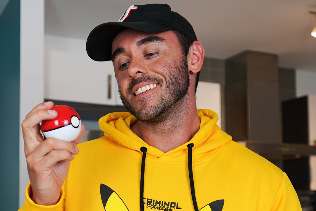 Corey stands in a bright yellow sweater holding a pokemon ball in his hand while smiling and looking at it