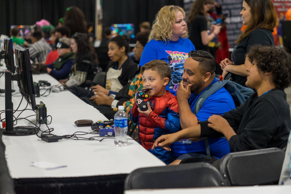 A kid in a spiderman cosplay sitting on his dad's lap while playing with a video game console