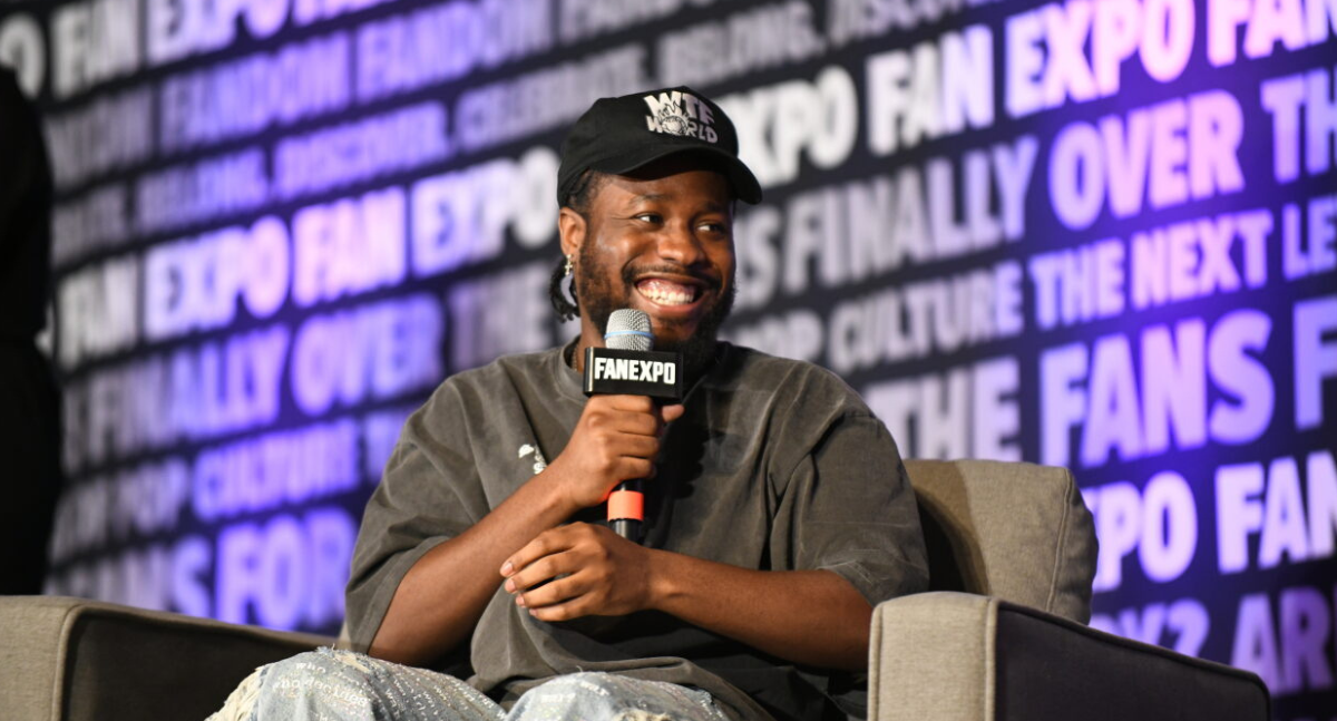 A very happy Shameik Moore smiles as the moderator speaks, holding his microphone just below his chin
