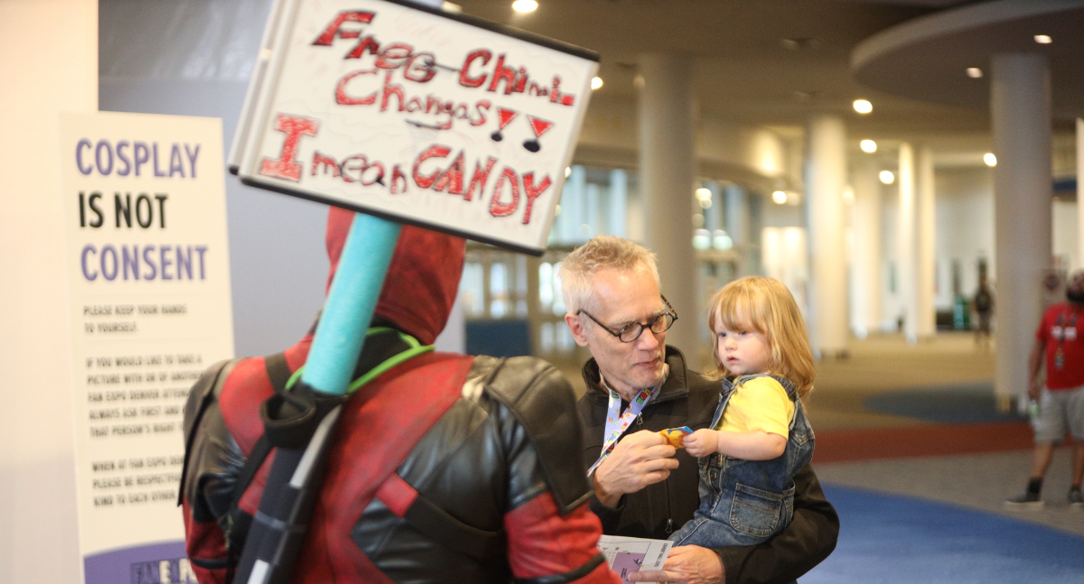 Deadpool hands out free candy to attendees at FAN EXPO. A father and daughter duo approach and take some swedish fish. She is unsure how she feels.