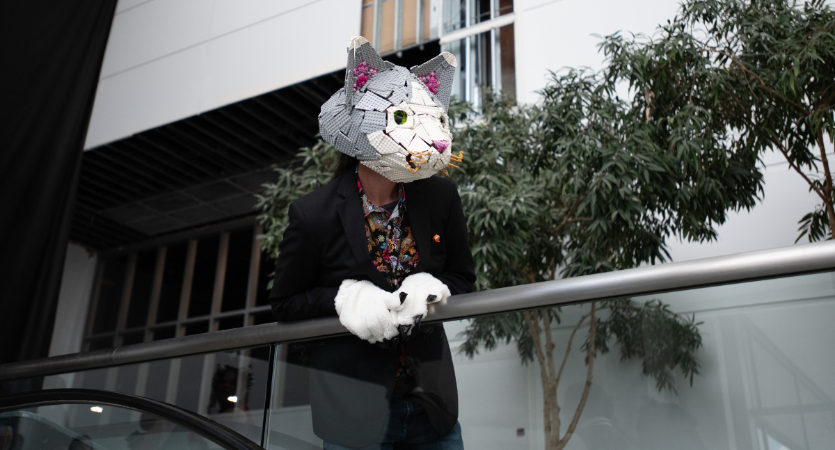 A cosplayer stands at the top of the escalator - their head is a cat head completely made out of lego, and they are wearing soft gloves. the head is grey and white lego, with pink inner ears
