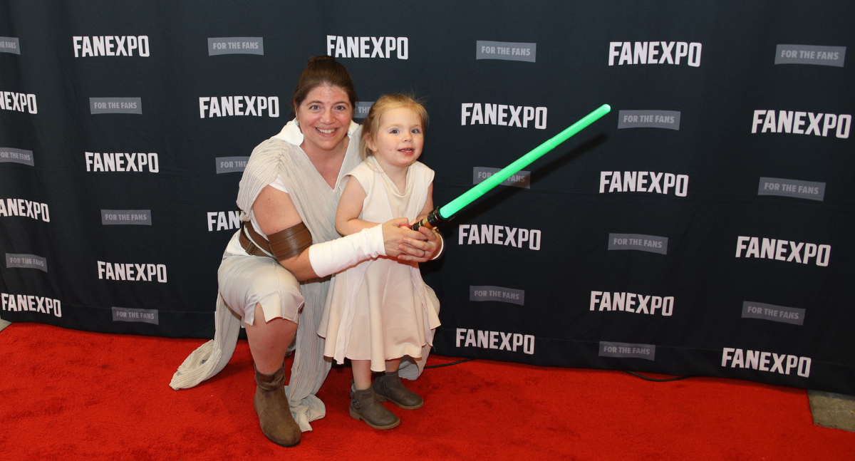 Cosplay Red Carpet - Mom and daughter are both dressed up as Ray, Mom is helping toddler hold green lightsaber
