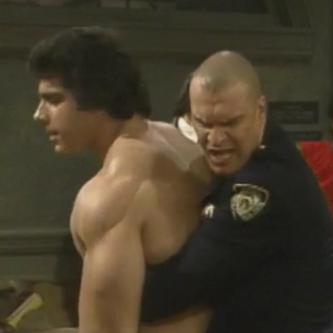 Lou is shirtless and is held by the bald security guard. Off screen is a little old woman trying to hit Lou.