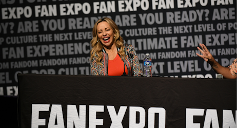 Tara Strong shares a laugh with the audience. She smiles wide in the foreground as the moderator, out of frame, right, speaks - his hands visible from right side.