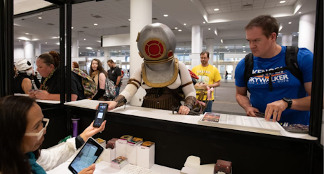 A couple comes to the ticketing counter to collection their tickets. The staff takes a phone displaying a QR code from a cosplayer outfitted with a deep sea diver's helmet