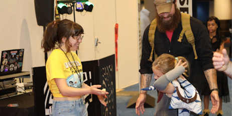 A young fan, dressed as Ahsoka Tano, collects her winnings from our FAN Fashion Catwalk raffle with her dad. The crew member giving her the prize smiles as the young girl accepts