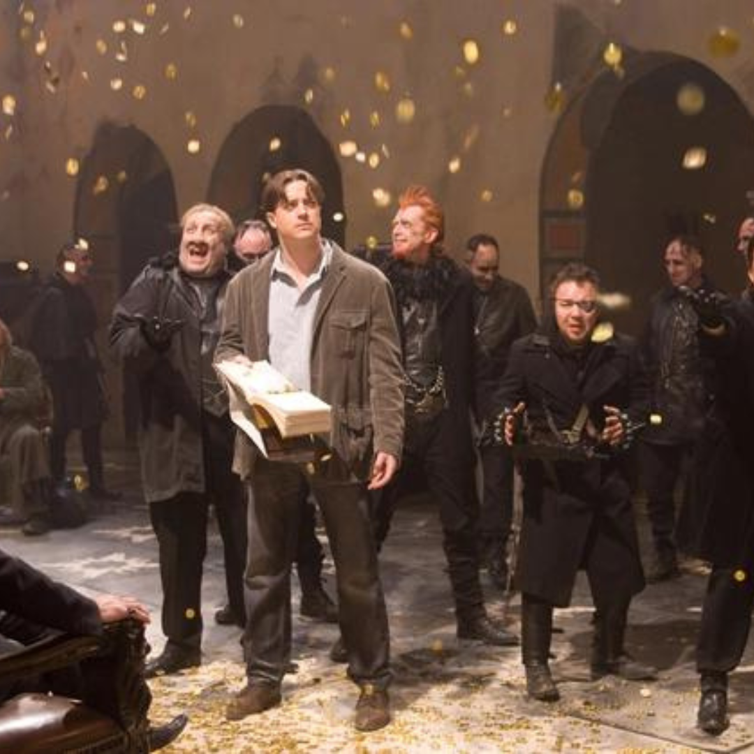 In an old room with arch doorways, Brendan Fraser's character is holding a book looking disappointed as coins fall from the sky. The scary and intense men surrounding him are ecstatic for the gold.