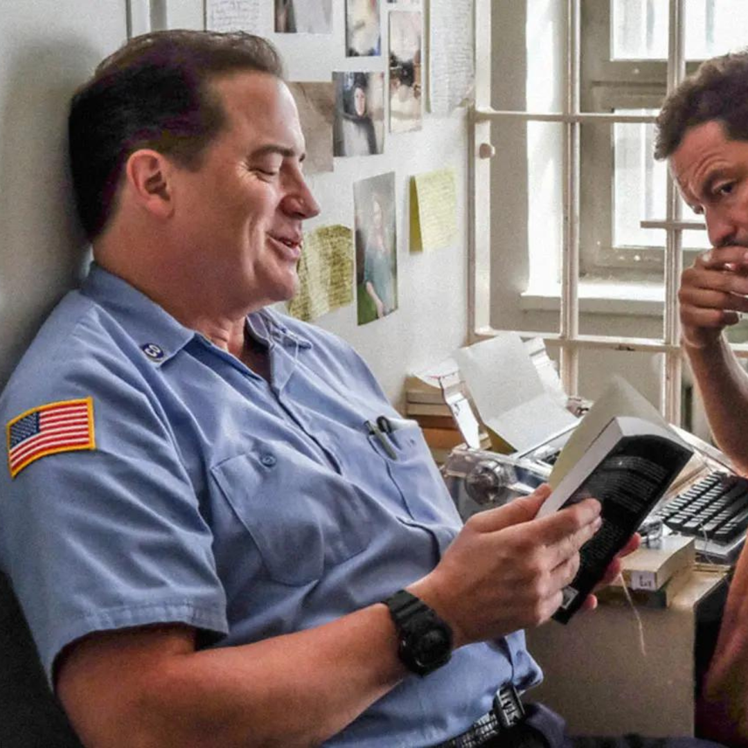 Brendan Fraser sitting in a jail is a jailer's uniform as he is smiling and reading a book next to an inmate typing on a type writer