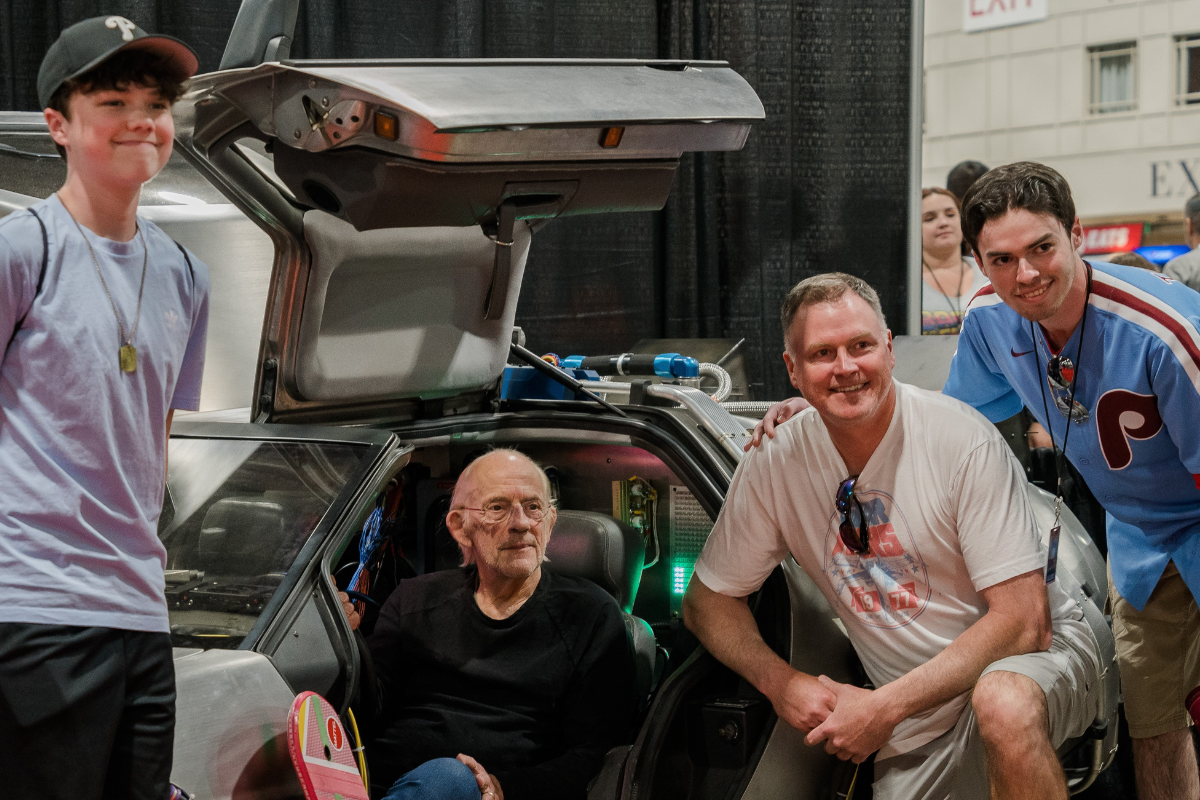 Christopher Lloyd sitting in a DeLorean with 3 guys posing next to him