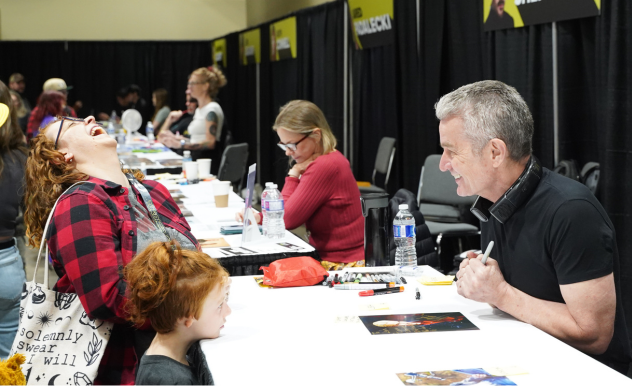 James Marsters smiles and laughs with a fan. Her young child, standing next to her, rests her chin on the table