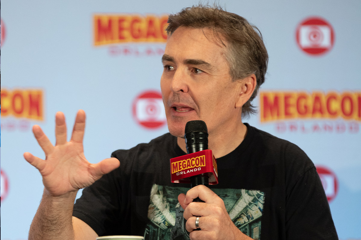 Nolan North speaks with his hand up and other hand holding a microphone