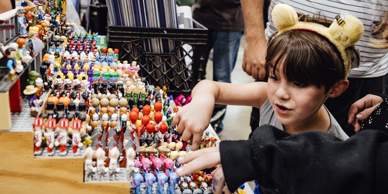 A young boy wearing a bear headband excitedly looks at a vendor’s display filled with colorful figurines and toys during a convention.