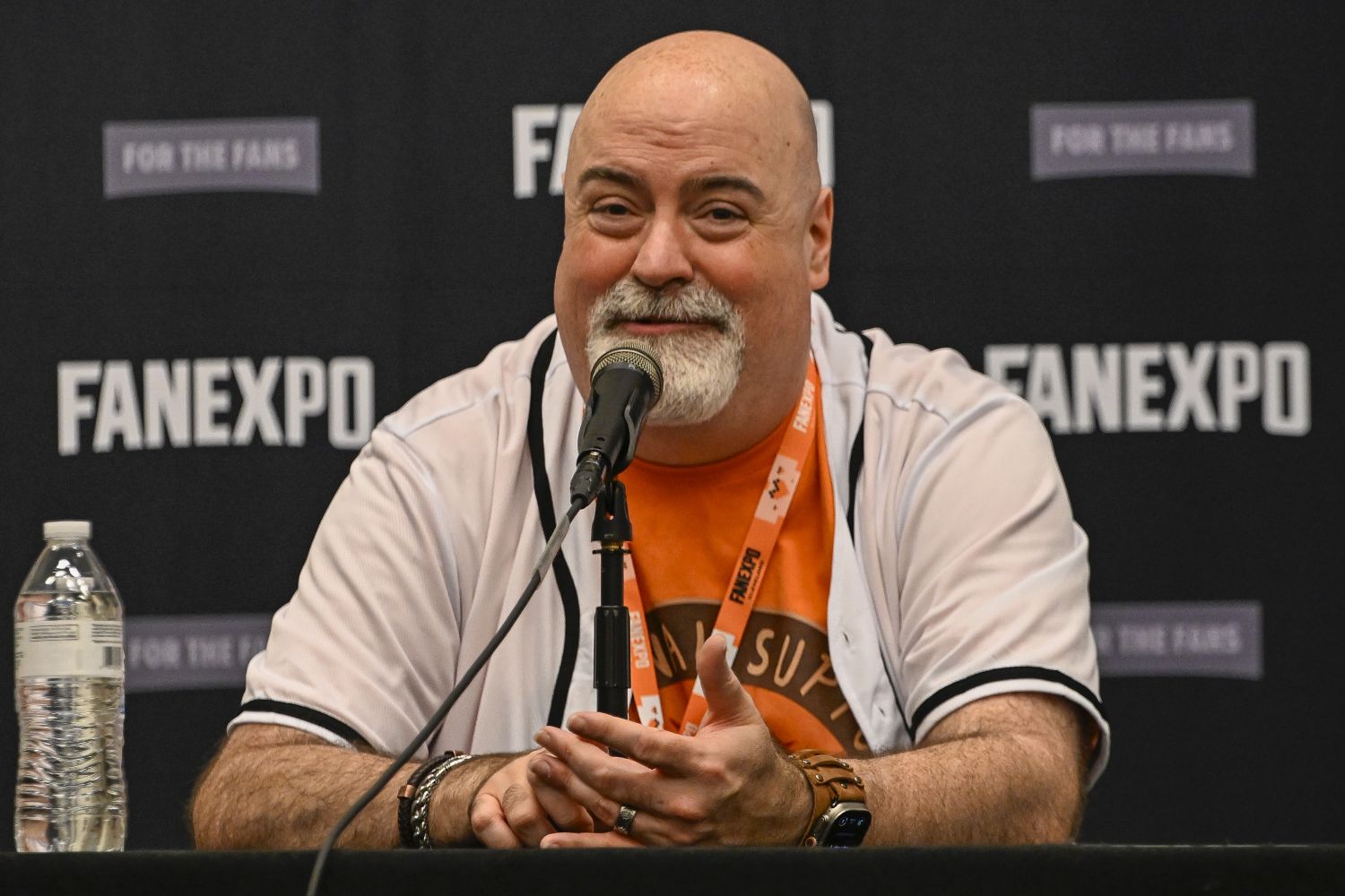 Kyle Hebert, with a white goatee and kind expression, speaks into a microphone at a FAN EXPO panel. He wears a white baseball-style jersey over an orange shirt with a FAN EXPO lanyard around his neck. He gestures slightly with his hands as he speaks, and a water bottle sits on the table beside him.