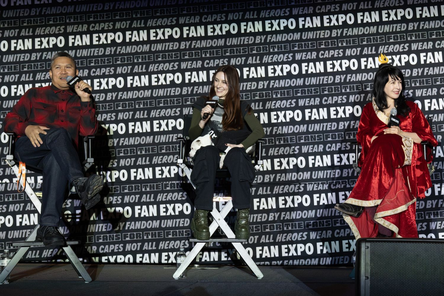 Dante Basco, Michaela Jill Murphy, and Grey DeLisle sit in director’s chairs on stage at FAN EXPO. On the left, Dante is wearing a red-and-black plaid button-down shirt and dark jeans and speaks into a microphone. In the center, Michaela has long, straight brown hair, holds a black-and-white dog on her lap, wearing a green long-sleeve shirt and dark cargo pants with olive green platform boots. On the right, Grey DeLisle is in a flowing red-and-gold fantasy-style costume, smiling at the others. The FAN EXPO step-and-repeat banner fills the background behind them.