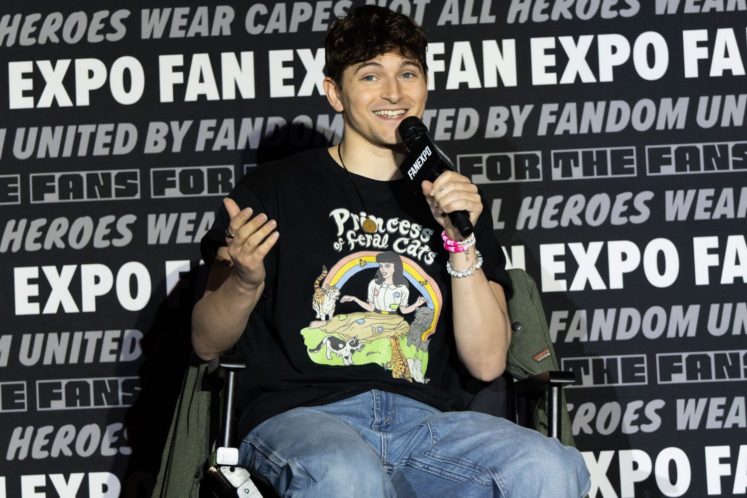 Blake Roman, with short, tousled brown hair sits in a director’s chair, smiling brightly and holding a FAN EXPO microphone in his right hand while gesturing with his left. He wears a black graphic t-shirt with the words “Princess of Feral Cats” and a colorful cartoon design, along with light blue jeans. He’s accessorized with several colorful beaded bracelets. The FAN EXPO step-and-repeat backdrop reads “UNITED BY FANDOM” and “NOT ALL HEROES WEAR CAPES” in various styles behind him.