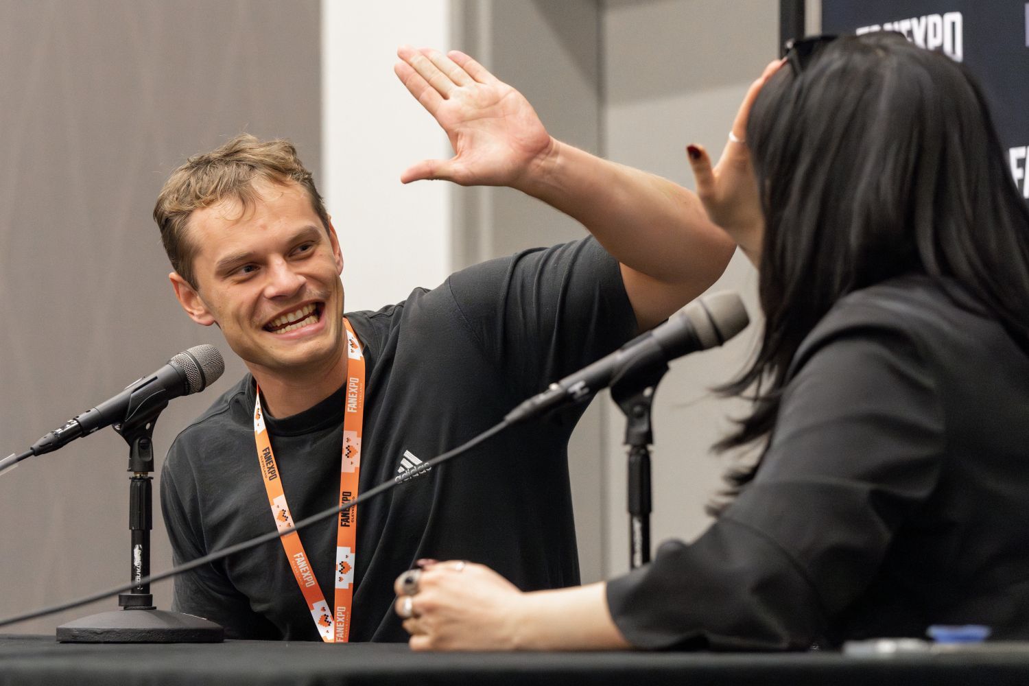 Aidan Scott, with short, light brown hair enthusiastically reaches out to high-five a woman seated across from him. He’s grinning wide, wearing a black Adidas shirt and a bright orange FAN EXPO lanyard. The woman, partially turned away from the camera, has long black hair and wears black sunglasses pushed up on her head along with a black outfit. They're sitting behind microphones at a panel table, mid-laugh.
