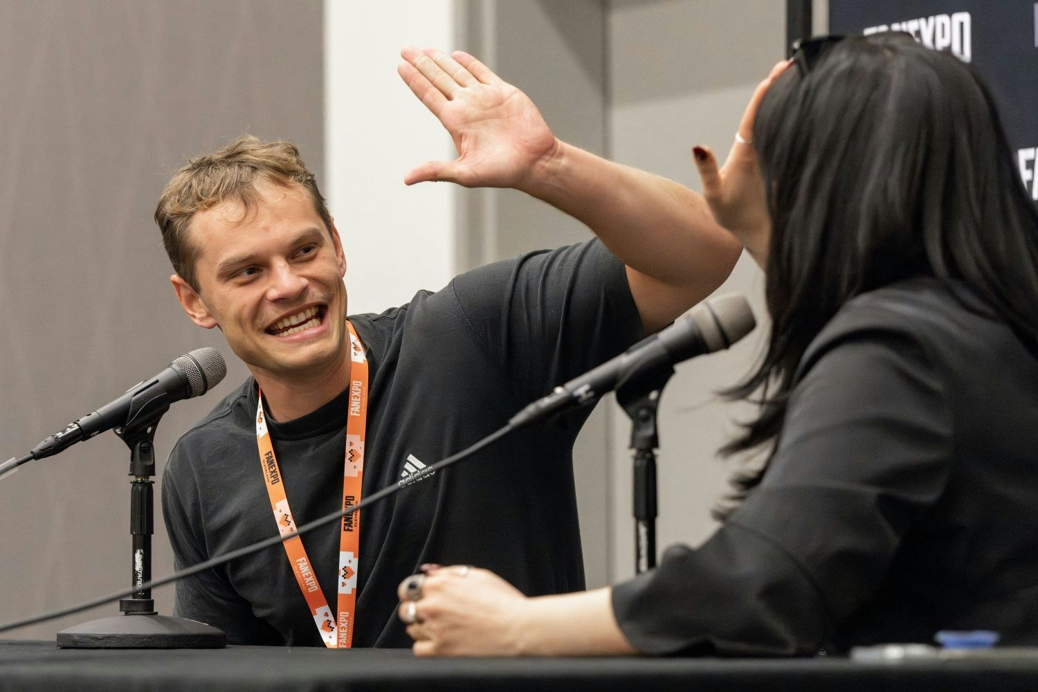 Aidan Scott, with short, light brown hair enthusiastically reaches out to high-five a woman seated across from him. He’s grinning wide, wearing a black Adidas shirt and a bright orange FAN EXPO lanyard. The woman, partially turned away from the camera, has long black hair and wears black sunglasses pushed up on her head along with a black outfit. They're sitting behind microphones at a panel table, mid-laugh.