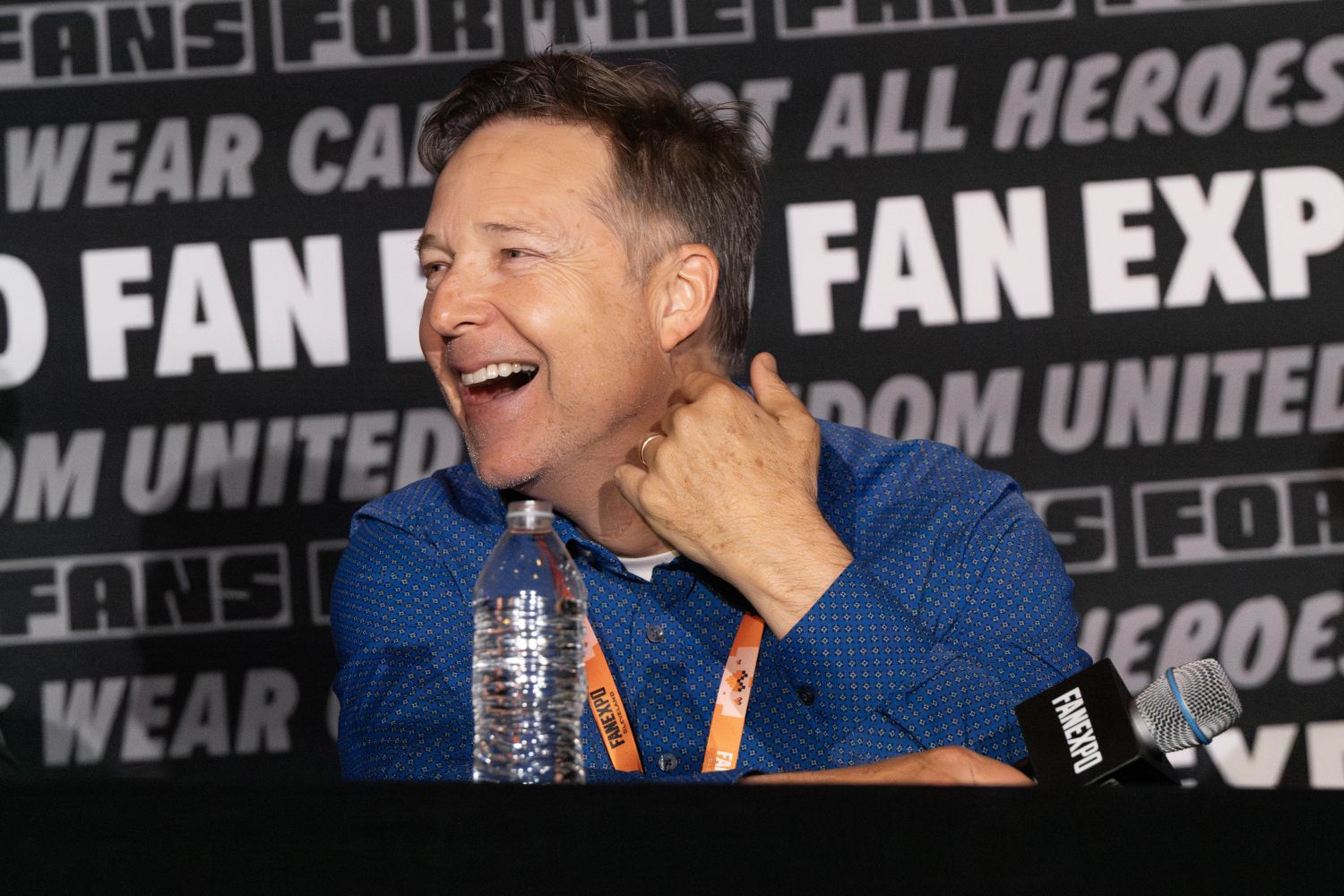 George Newbern, with short, graying hair and a bright smile laughs while turning to his left. He’s seated at a panel table, wearing a royal blue, patterned button-down shirt and an orange FAN EXPO lanyard. A half-full water bottle and a FAN EXPO mic rest on the table in front of him. The background features a repeating FAN EXP