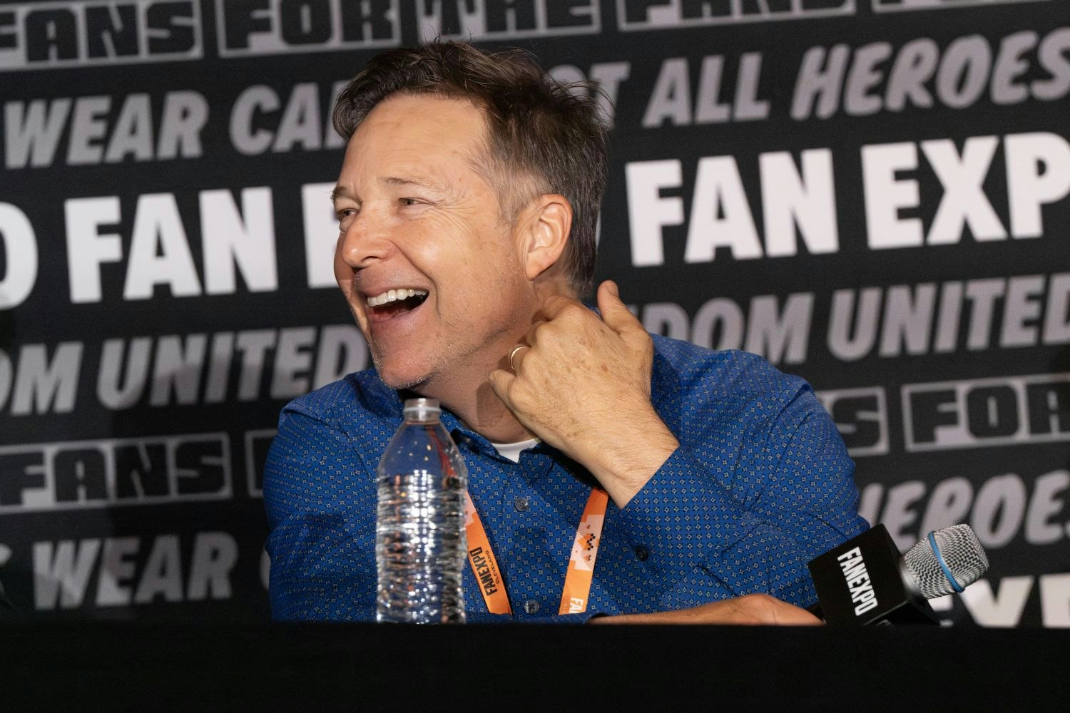 George Newbern, with short, graying hair and a bright smile laughs while turning to his left. He’s seated at a panel table, wearing a royal blue, patterned button-down shirt and an orange FAN EXPO lanyard. A half-full water bottle and a FAN EXPO mic rest on the table in front of him. The background features a repeating FAN EXP