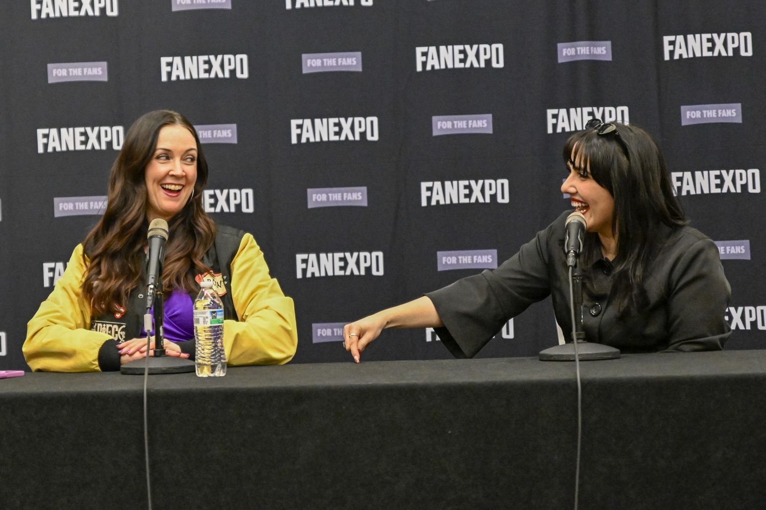 Stephanie Young and a female panelist sit at a panel table mid-laugh. On the left is Stephanie Young with long, wavy brown hair wears a gold and black varsity-style jacket over a purple top, sitting behind a microphone and water bottle. On the right, the female panelist has jet-black hair styled with bangs and sunglasses pushed up on her head leans over dramatically with a big smile, wearing a black outfit. They sit in front of a FAN EXPO branded wall.