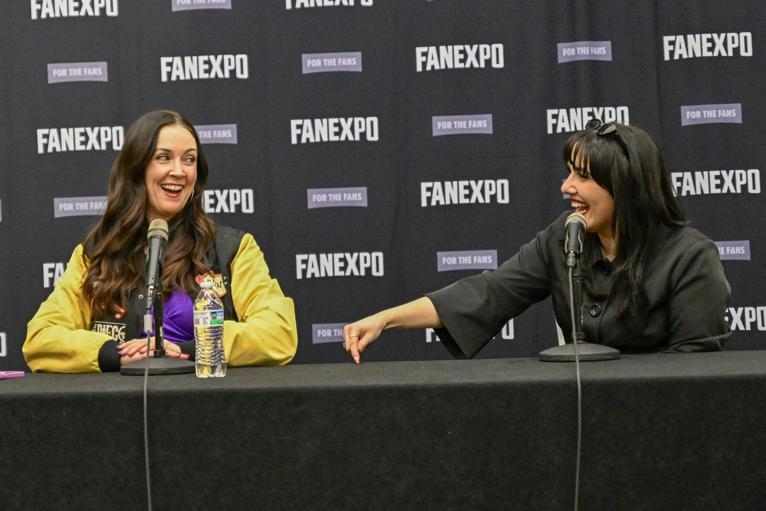 Stephanie Young and a female panelist sit at a panel table mid-laugh. On the left is Stephanie Young with long, wavy brown hair wears a gold and black varsity-style jacket over a purple top, sitting behind a microphone and water bottle. On the right, the female panelist has jet-black hair styled with bangs and sunglasses pushed up on her head leans over dramatically with a big smile, wearing a black outfit. They sit in front of a FAN EXPO branded wall.