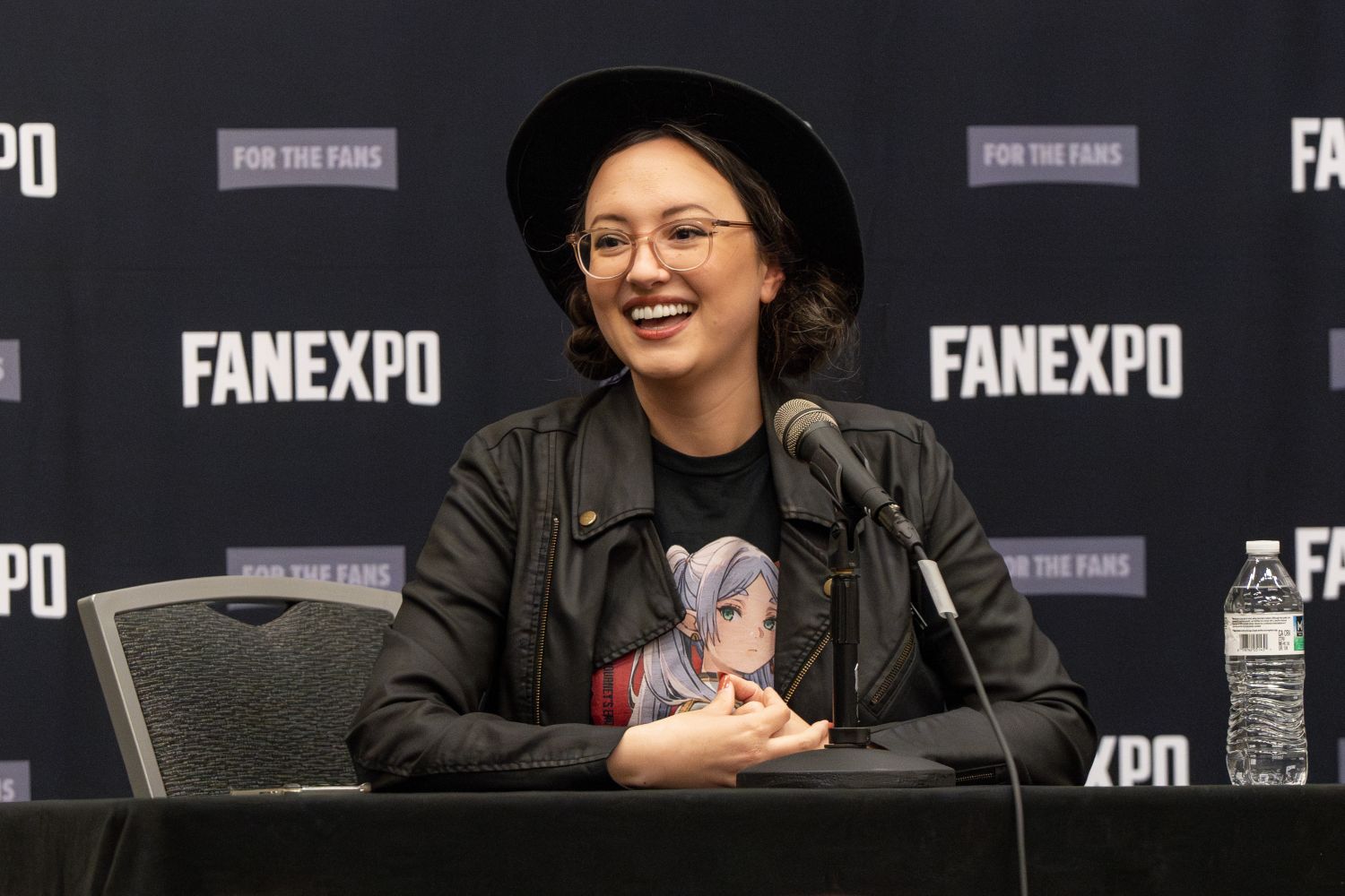 Mallorie Rodak, wearing a black wide-brimmed hat and round glasses smiles enthusiastically while speaking into a microphone. She wears a black faux leather jacket over a black shirt with an anime-style character on the front. Her hair is pulled back into two low buns. She sits at a panel table in front of a FAN EXPO step-and-repeat backdrop.