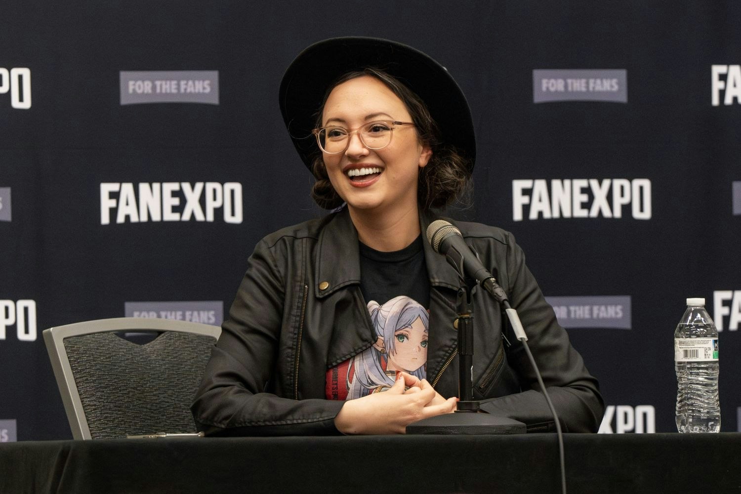 Mallorie Rodak, wearing a black wide-brimmed hat and round glasses smiles enthusiastically while speaking into a microphone. She wears a black faux leather jacket over a black shirt with an anime-style character on the front. Her hair is pulled back into two low buns. She sits at a panel table in front of a FAN EXPO step-and-repeat backdrop.