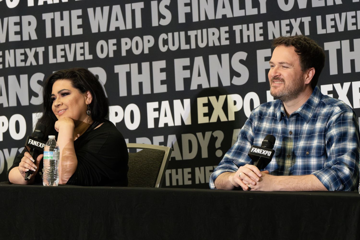 Leah Clark , with long, black hair styled in glamorous waves and bold makeup sits besides Chris Wehkamp  in a plaid button-down shirt. Both are smiling warmly while holding FAN EXPO microphones at a panel table. Leah wears a black top and hoop earrings and leans slightly on her hand. The background is covered with bold phrases like “THE WAIT IS FINALLY OVER” and “THE NEXT LEVEL OF POP CULTURE.”