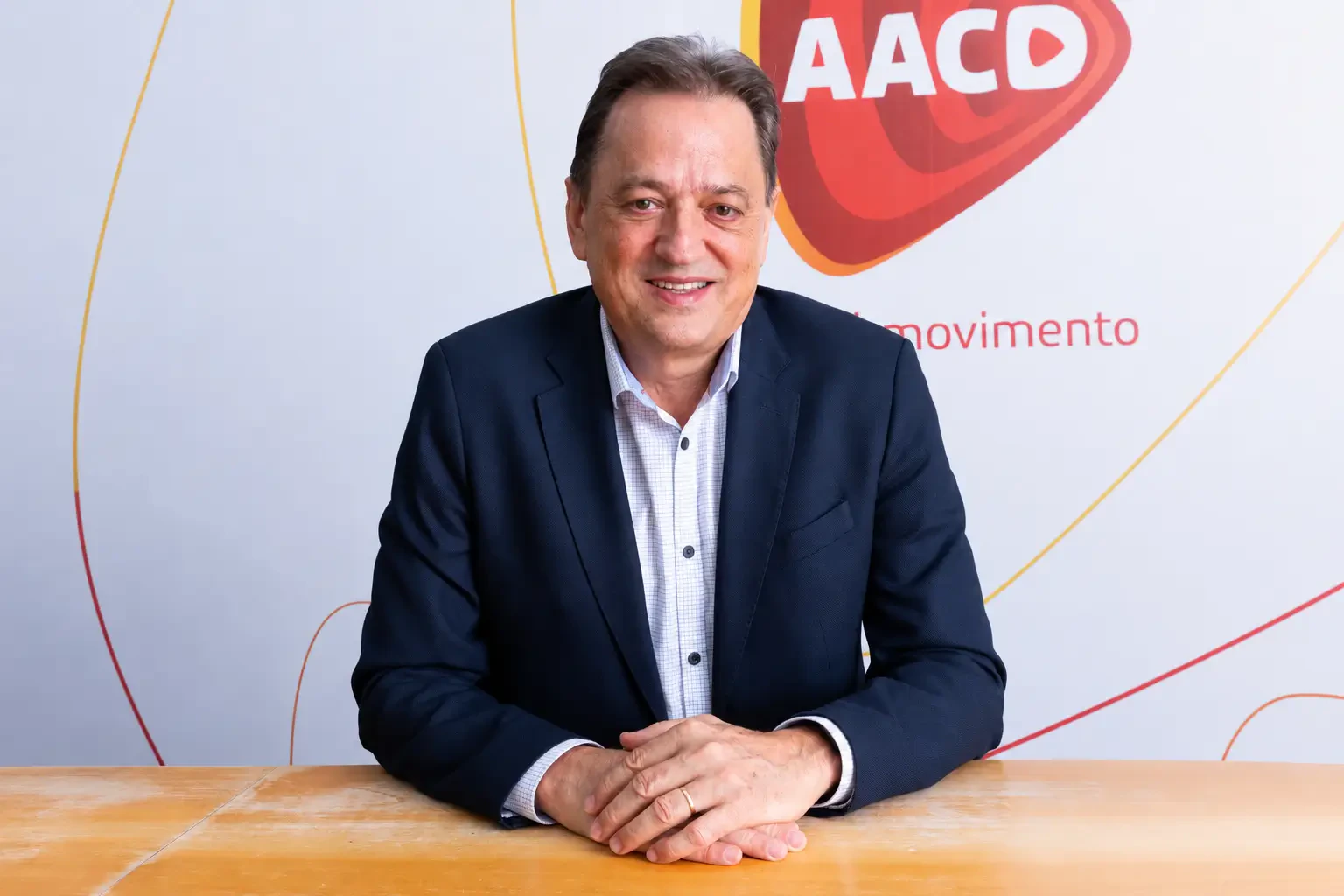 Valdesir Galvan sorrindo sentado à mesa de paletó azul-marinho, com painel de fundo da AACD.