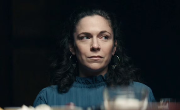 A woman in a blue blouse, with curly brown hair looks to her right to a person off screen. She is clearly sitting at a dinner table surrounded by glassware