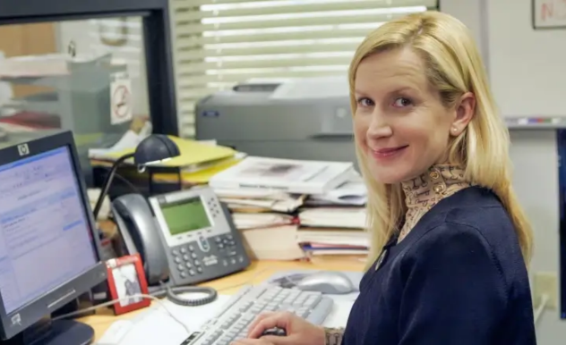 Angela turns her head to the side and smiles at the camera. She is typing in a document at her desk. 