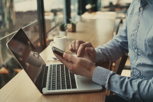Photograph of a person in a blue button up shirt sitting at a cafe bar with a cup of coffee in front of a laptop looking at their phone.