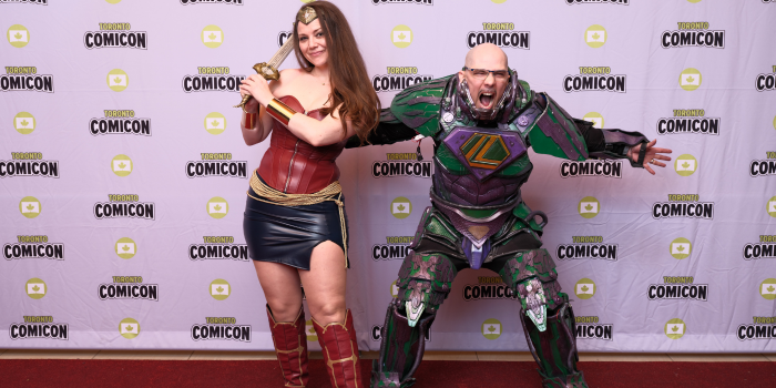 A female and male cosplayer pose on the carpet together. She swings her sword behind her head, and he stands, mouth agape in a yell, screaming toward the camera.