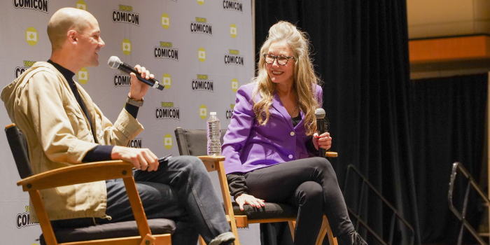 Left to right: Moderator and Mary McDonnell. They are seated in front of a Toronto Comicon branded backdrop. Both with their legs crossed, with big smiles