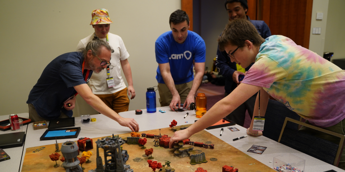 A group of individuals play battlefront together in the Focht's Network's gaming lounge. three of five individuals are placing items on the play field including miniature figures and cards.